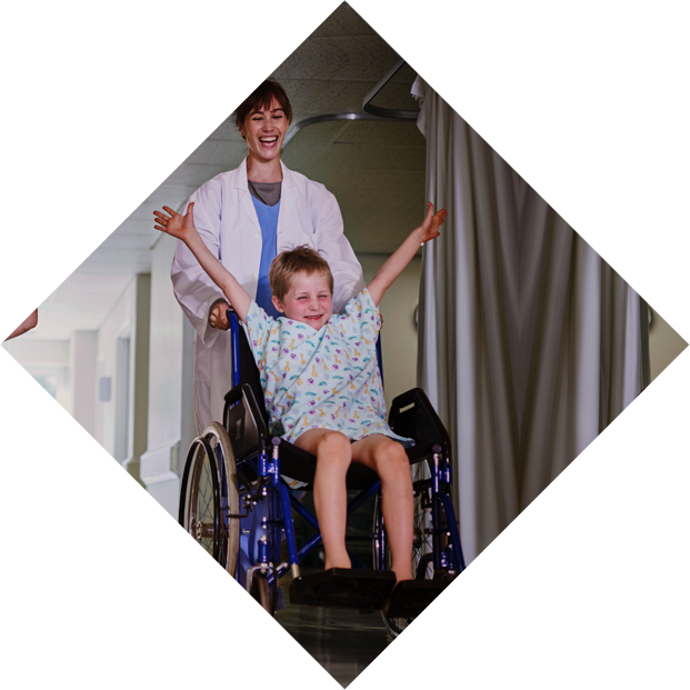 Zwei Kinder werden mit Rollstühlen über einen Krankenhausflur geschoben. Die Kinder strecken die Arme in die Luft und haben Spaß.